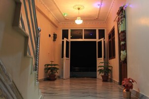 Um corredor com piso de mármore, duas plantas em vasos, um candelabro e uma porta que dá para uma varanda.