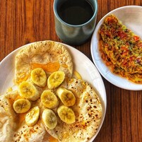Um prato de crepes com fatias de banana e calda, uma tigela de bolinhos de legumes e uma xícara de café preto sobre uma mesa de madeira.