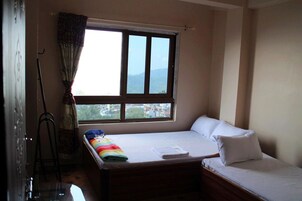 Um quarto pequeno com uma cama de solteiro, janela com cortinas e vista para a paisagem urbana.