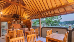An outdoor seating area with wooden tables and chairs, a bar with shelves of bottles, and a view of the surrounding landscape. - Kastapar Bridge Guest House (Lembongan Island)
