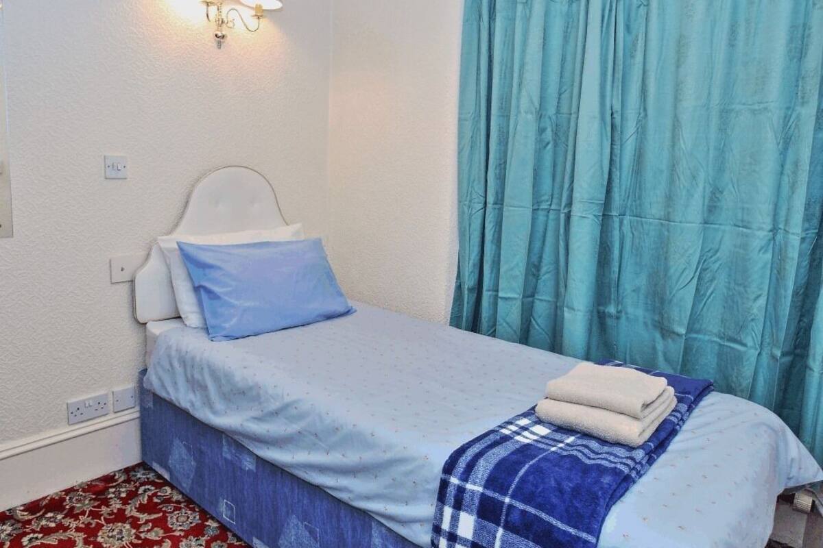 A single bed with a blue and white bedspread, a blue pillow, and a folded towel on the bed. There is a wall-mounted light fixture above the bed.