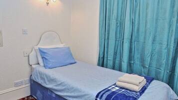 A single bed with a blue and white bedspread, a blue pillow, and a folded towel on the bed. There is a wall-mounted light fixture above the bed.