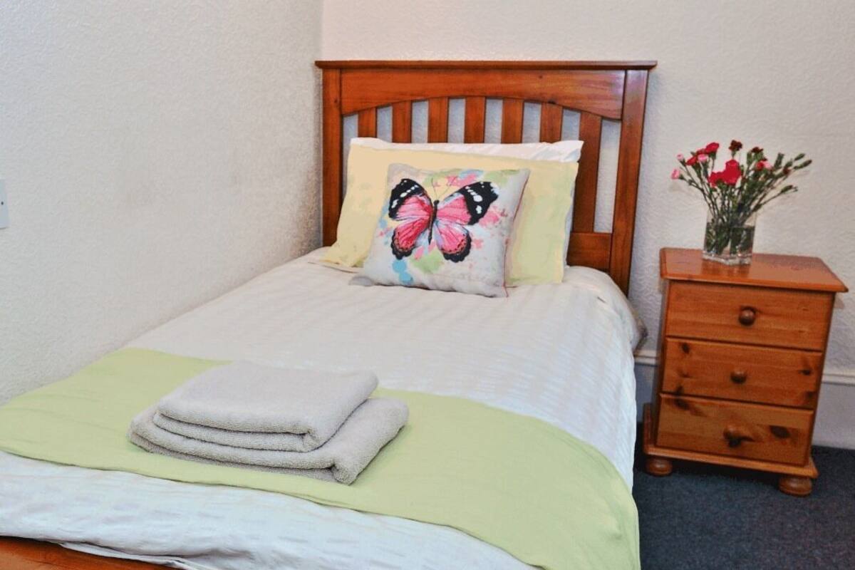 A neatly made bed with a green bedspread, a pillow with a butterfly design, and a folded towel. There is a wooden nightstand with a vase of flowers beside the bed.