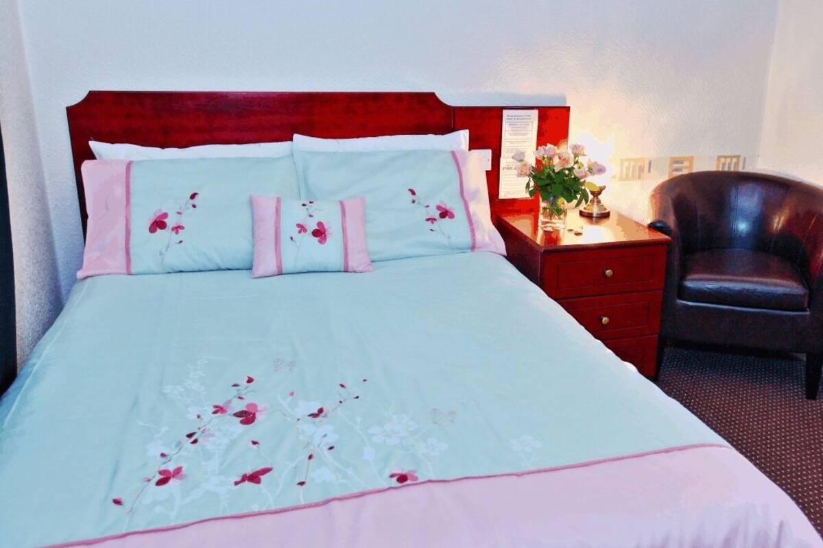 A neatly made bed with floral bedding, a wooden headboard, a bedside table with a lamp, and a leather chair.