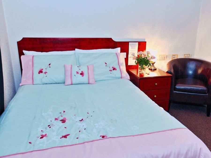A neatly made bed with floral bedding, a wooden headboard, a bedside table with a lamp, and a leather chair.