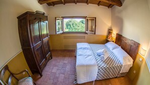 Une chambre avec une armoire en bois, un lit recouvert d’une housse à motifs à pois, une fenêtre donnant sur un paysage verdoyant et un plancher en briques.