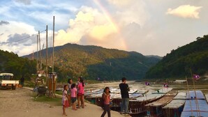 Margem de rio com barcos, um arco-íris e uma paisagem montanhosa.