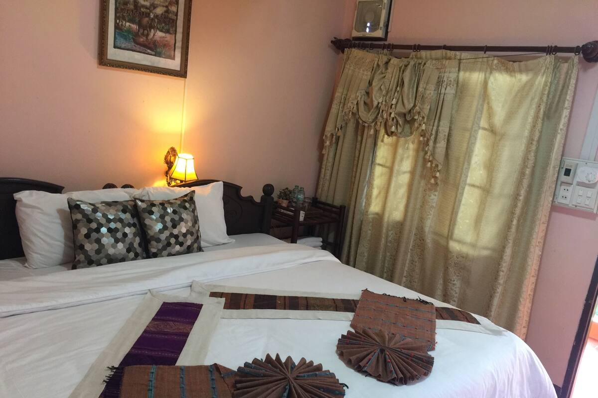 A bedroom with a bed, pillows, a lamp, curtains, and a wall-mounted air conditioner.
