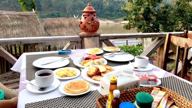 Mesa posta com itens de café da manhã, incluindo panquecas, ovos e torradas, com vista para um rio e colinas arborizadas.
