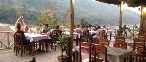 An outdoor dining area with tables and chairs, surrounded by potted plants and overlooking a mountainous landscape.