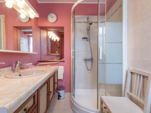A bathroom with a glass-enclosed shower, a double sink vanity, a mirror with lights, and a clock on the wall.