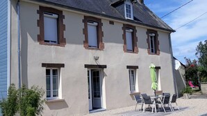 Ein zweistöckiges Gebäude mit einer beigen Fassade, mehreren Fenstern mit Fensterläden, einer Tür und einer kleinen Terrasse mit Tisch und Stühlen.
