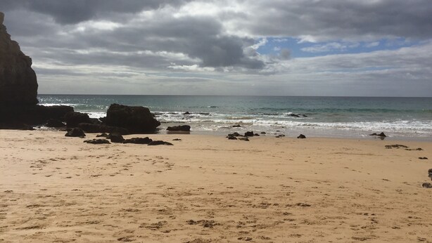 Uma praia com afloramentos rochosos e areal, sob um céu nublado.