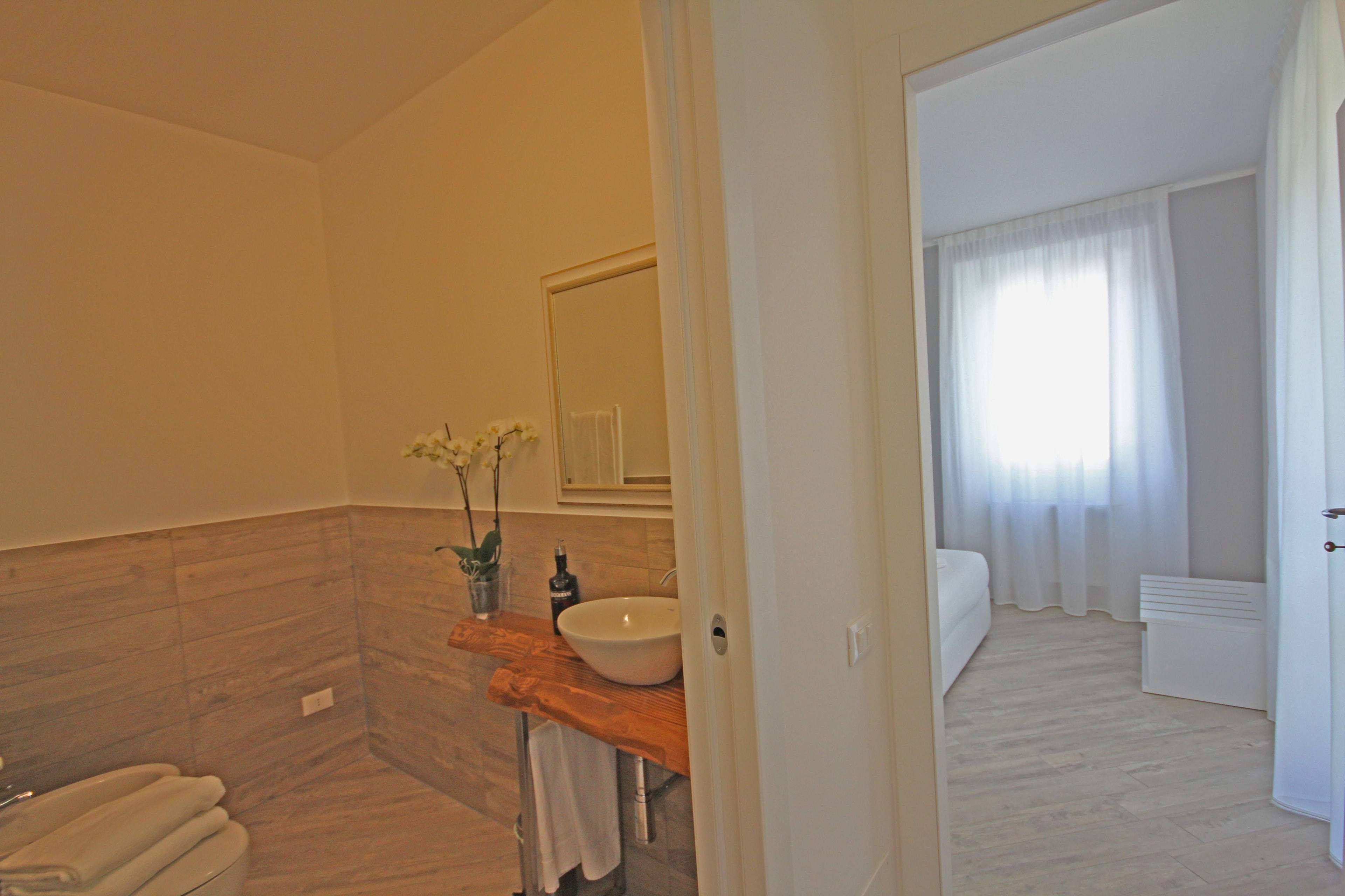 Un bagno con un mobile vanitario in legno, un lavandino bianco e uno specchio. Si vede il water con un portasciuga e una piccola pianta. Il pavimento e le pareti sono di colore chiaro.