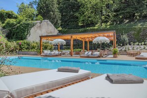 Un área de piscina con sillas de descanso y sombrillas, rodeada de vegetación y un muro de piedra.