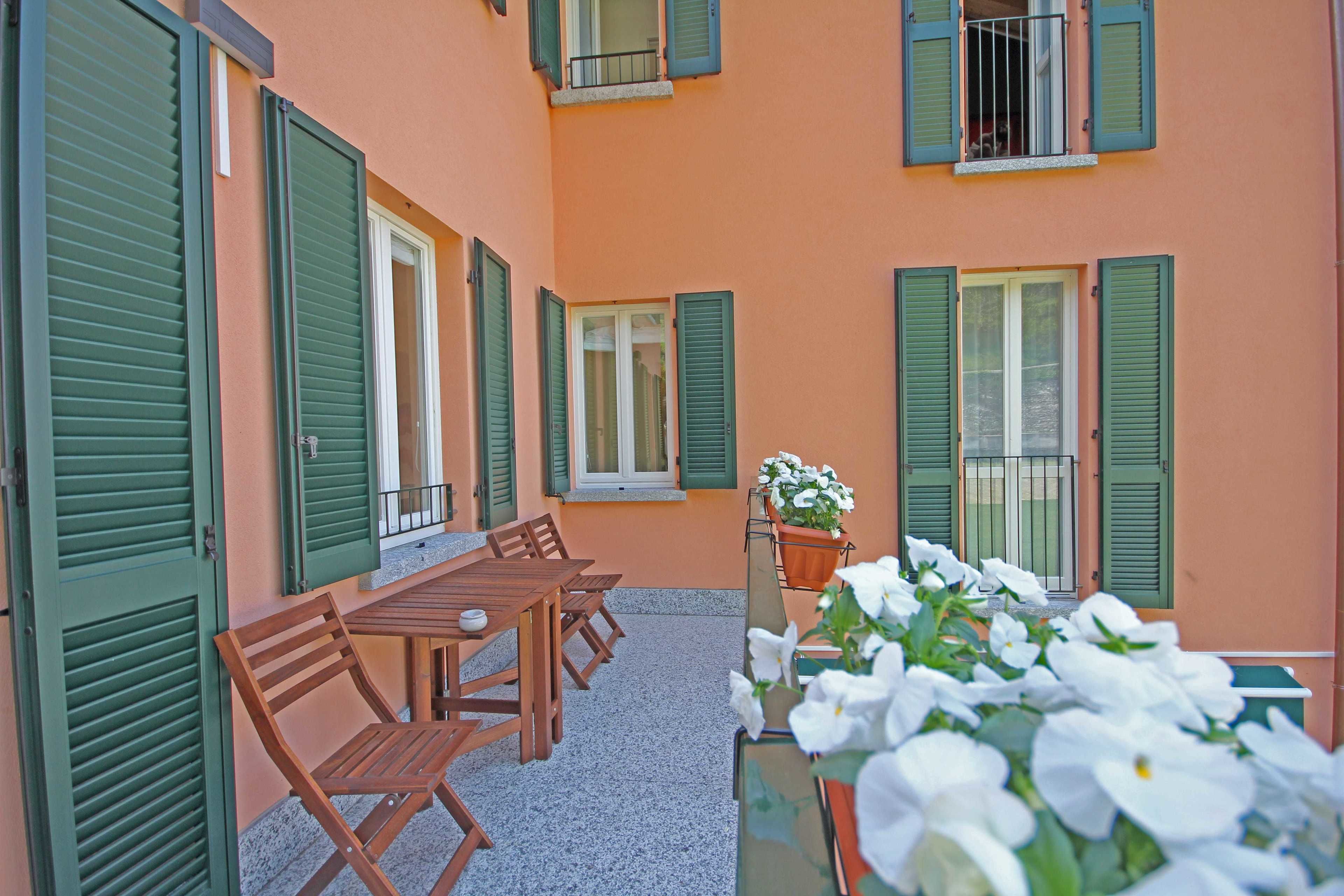 Un patio con una mesa y sillas de madera, una planta en maceta con flores blancas y un edificio con contraventanas verdes.