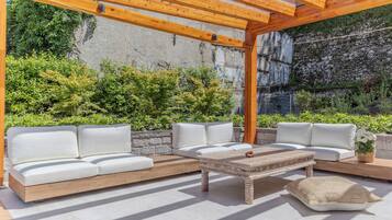 Un patio al aire libre con una pérgola de madera, asientos blancos acolchados y un muro de piedra como fondo.