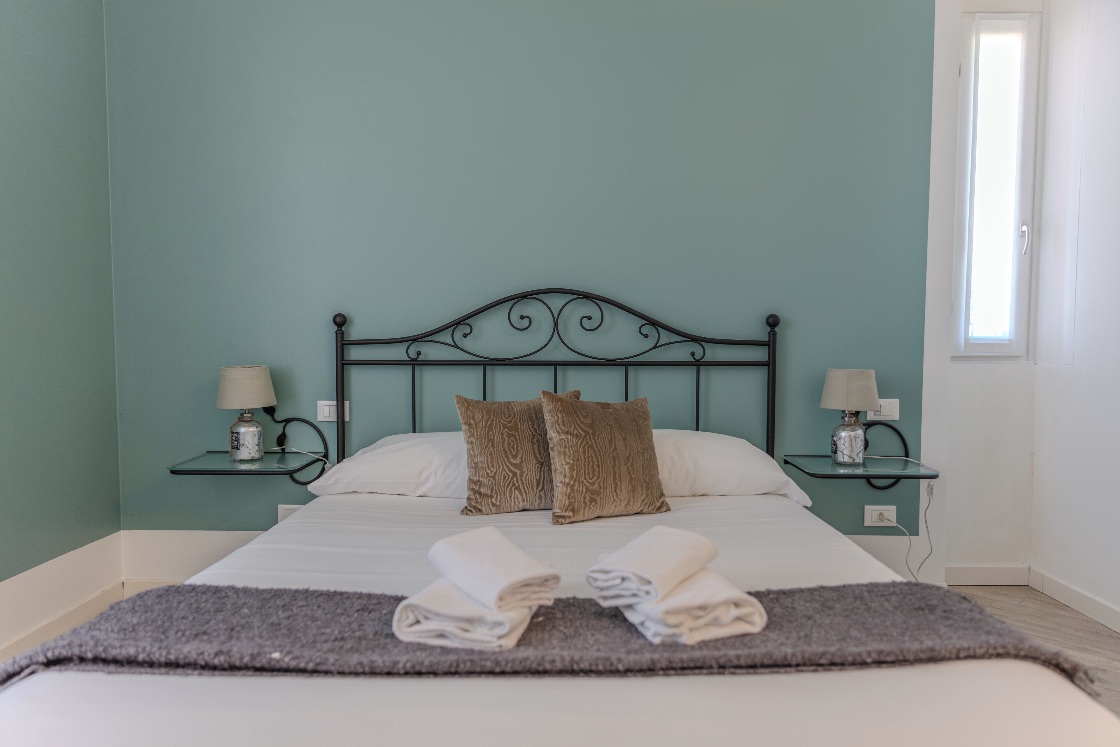 A neatly made bed with two pillows, a bedside table with a lamp, and a window with daylight.