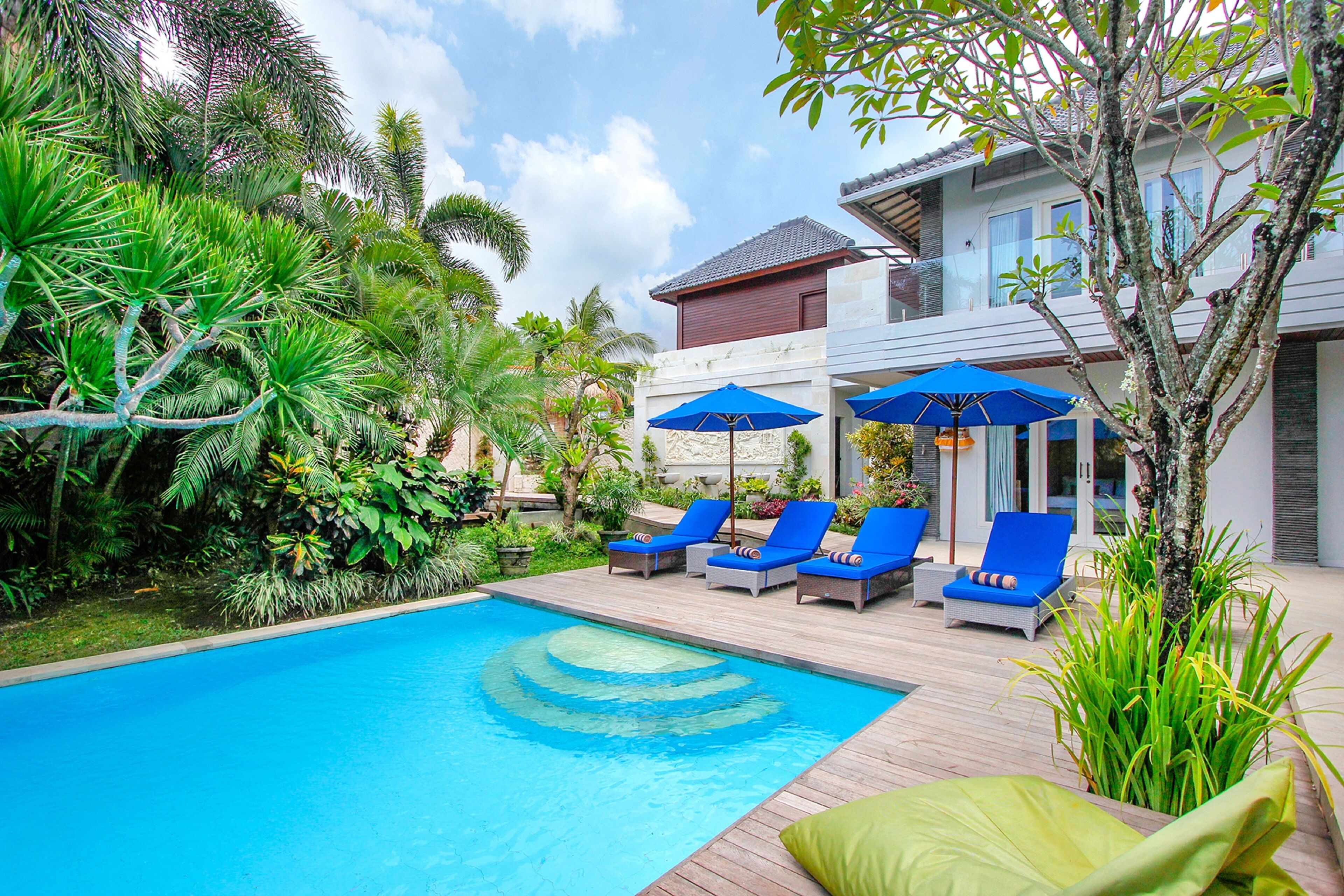A pool area with lounge chairs and umbrellas, surrounded by lush greenery and a modern building.