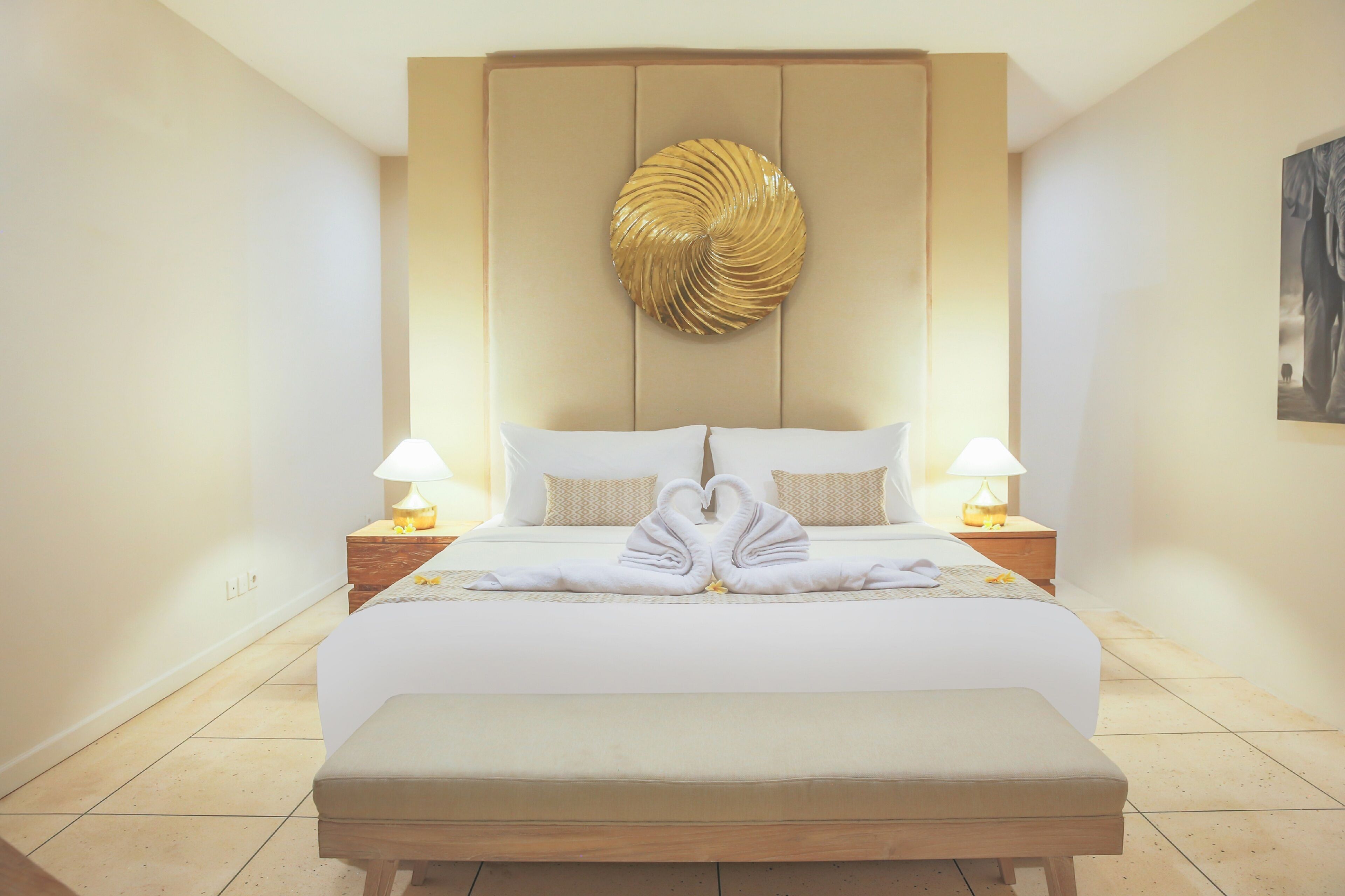 A hotel room with a large bed, two bedside tables with lamps, a wall-mounted decorative piece, and a bench at the foot of the bed.