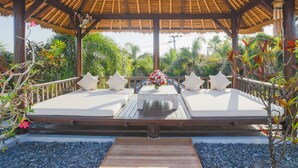 A wooden deck with two white cushioned seating areas, a central table with a floral arrangement, and a thatched roof structure. - Villa La Luvier (Canggu)