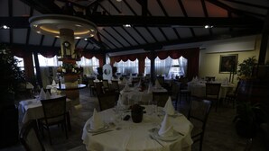 A dimly lit restaurant with round tables set for dining, featuring a central decorative column and a clock. - Guesthouse Tara (Belgrade)