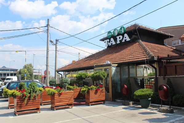 A building with a sign reading "TAPA" and a tiled roof. There are wooden planters with greenery and red flowers in front.