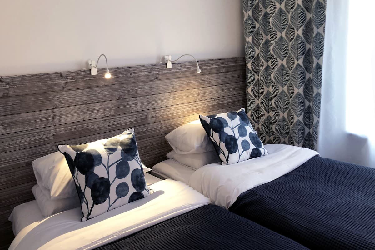 A double bed with white and dark blue bedding, two wall-mounted reading lights, and a patterned curtain.