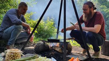 Two people cooking outdoors with a portable stove, surrounded by greenery.