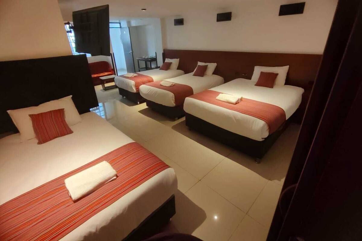 A hotel room with three beds, each with red and white bedding, and a separate seating area with a chair and table.