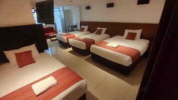 A hotel room with three beds, each with red and white bedding, and a separate seating area with a chair and table.