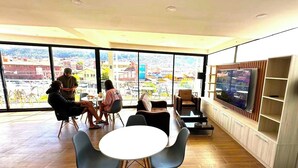 A spacious room with large windows offering a city view, a round table with chairs, and a television on the wall. - Mama's House (Cusco)