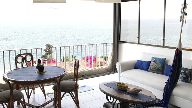 Een balkon met uitzicht op zee, een eettafel met stoelen, een bank en een hangplant.