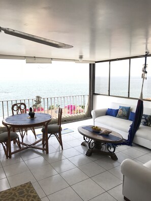Un balcón con vista al mar, una mesa de comedor con sillas, un sofá y una planta colgante.