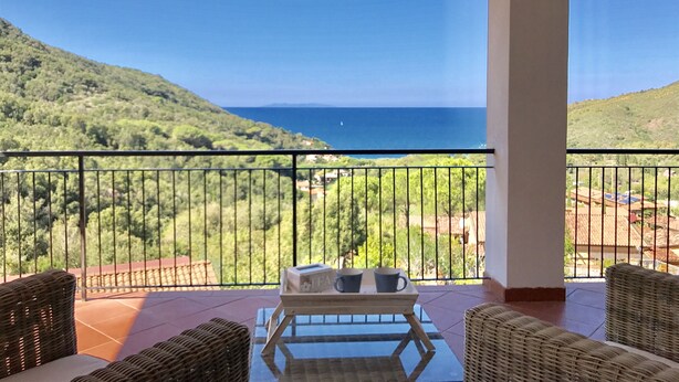 Un balcon agrémenté de meubles en osier, une table avec des tasses à thé et une vue sur la mer et les collines.
