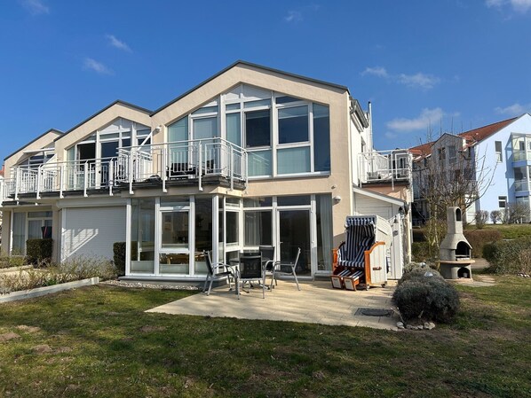 Une maison moderne à deux étages, dotée de grandes fenêtres, d’un balcon et d’un patio aménagé avec du mobilier d’extérieur.