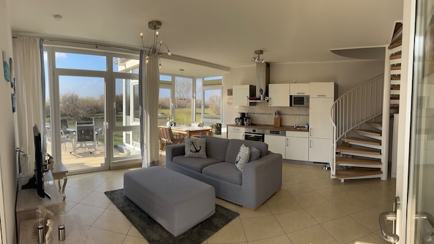 A spacious living area with a grey sofa and ottoman, a kitchen with white cabinets, and a staircase leading to an upper floor. - Haffnest with wonderful views of the Salzhaff  (Rerik)