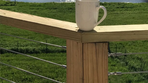Een witte mok op een houten leuning met uitzicht op een meer en een bebost heuvel in de verte.