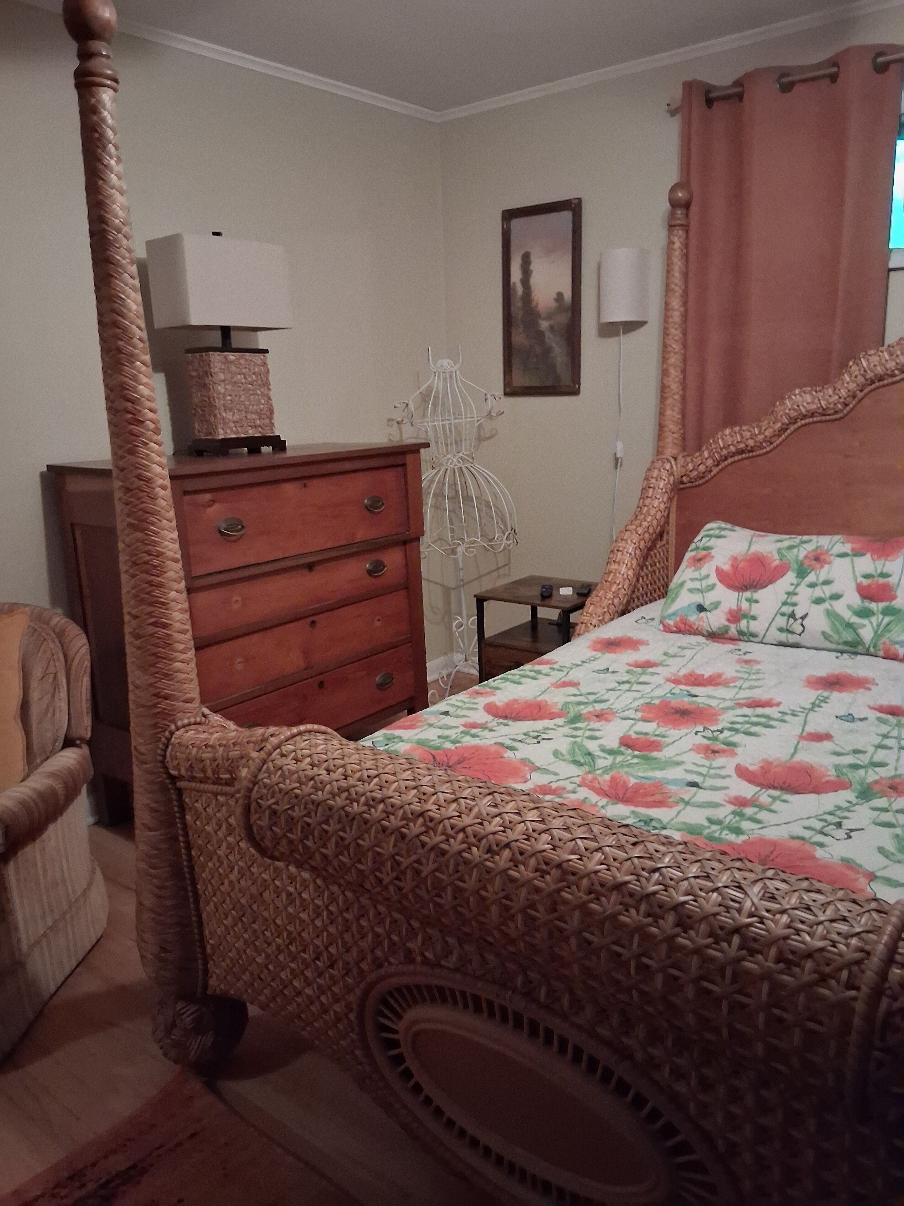 Una cama con dosel adornada con una colcha floral, un aparador de madera con cajones, una jaula blanca para pájaros y una pequeña mesa con lámpara.