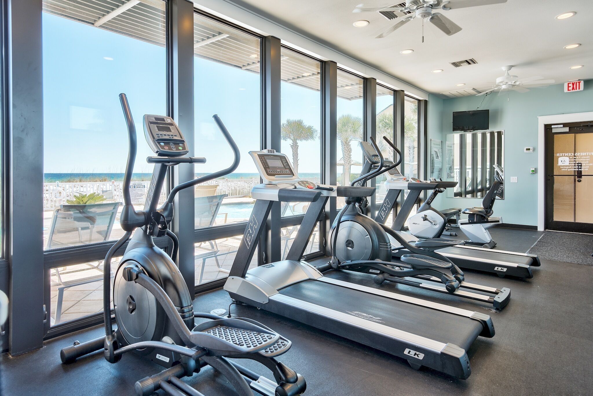 Un gimnasio con cintas de correr y elípticas, amplias ventanas con vista al océano y una señal de salida.