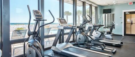 Un gimnasio con cintas de correr y elípticas, amplias ventanas con vista al océano y una señal de salida.