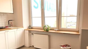 Een keuken met witte kastjes, een raam met uitzicht, een radiator en een klein krukje met een tijdschrift.