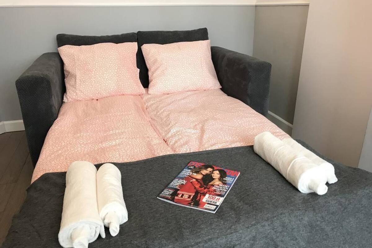 Ein Bett mit dunkelgrauem Kopfteil und zwei rosafarbenen Kissen. Auf dem Bett liegen zwei aufgerollte Handtücher, und ein Magazin ist dort platziert.