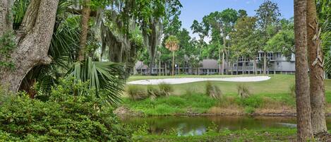 A golf course with a pond, surrounded by trees and a building in the background.