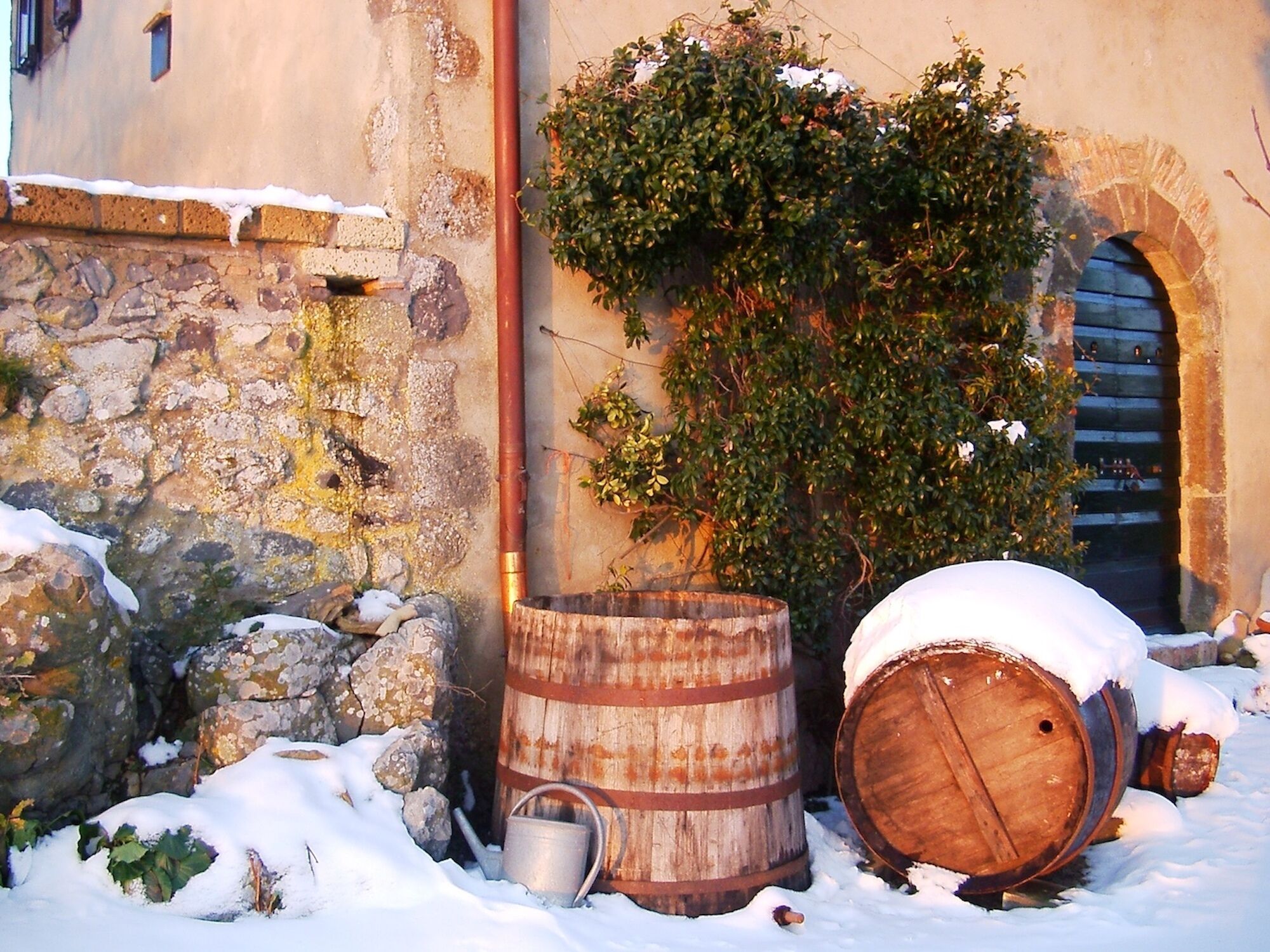 A rustic outdoor setting with a stone wall, wooden barrels, and a metal watering can in the snow.
