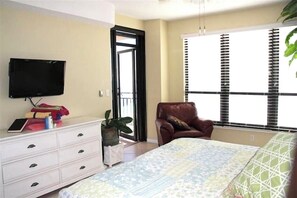 A bedroom with a bed, a TV mounted on the wall, a chair, a dresser, and a window with blinds. - SeaChase 602 East (Orange Beach)