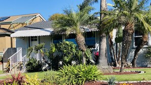 A residential house with a sloped roof, a front porch, and a well-maintained garden with various plants and a palm tree. - Quaint and Charming 2 bd, 1 bath,  Central Location, easily accessible! (San Diego)
