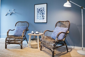 Two wicker chairs with patterned cushions, a small table with a woven basket, and a framed abstract art piece on a blue wall. - Johanna's Hotel (Bua)