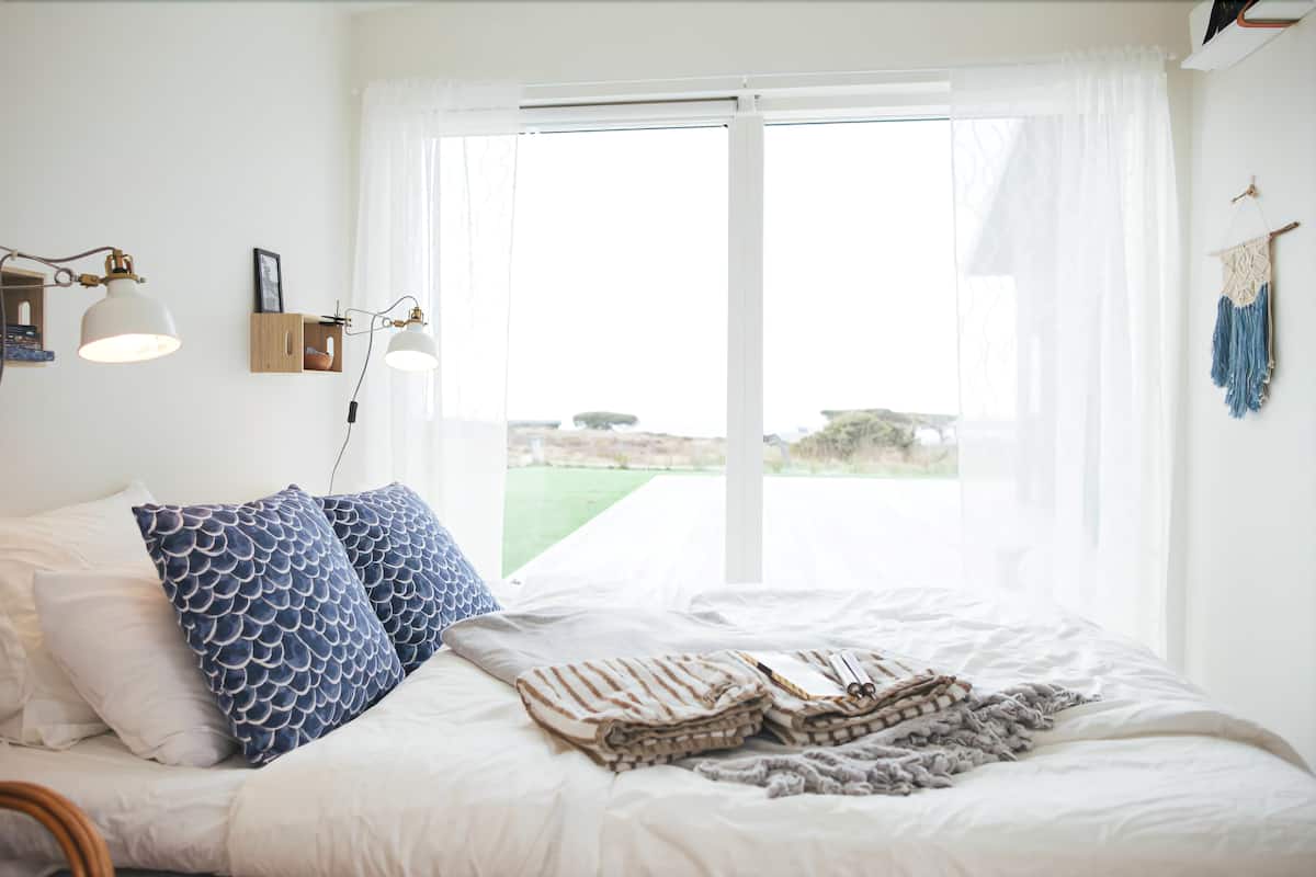 A bedroom with a bed, pillows, a bedside lamp, a window with curtains, and a view of the outdoors.