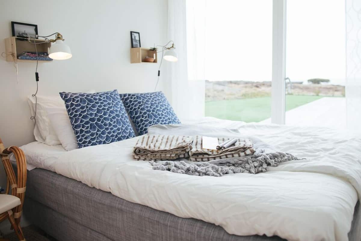 A neatly made bed with patterned pillows, a bedside table with a lamp, and a view of the outdoors through large windows.